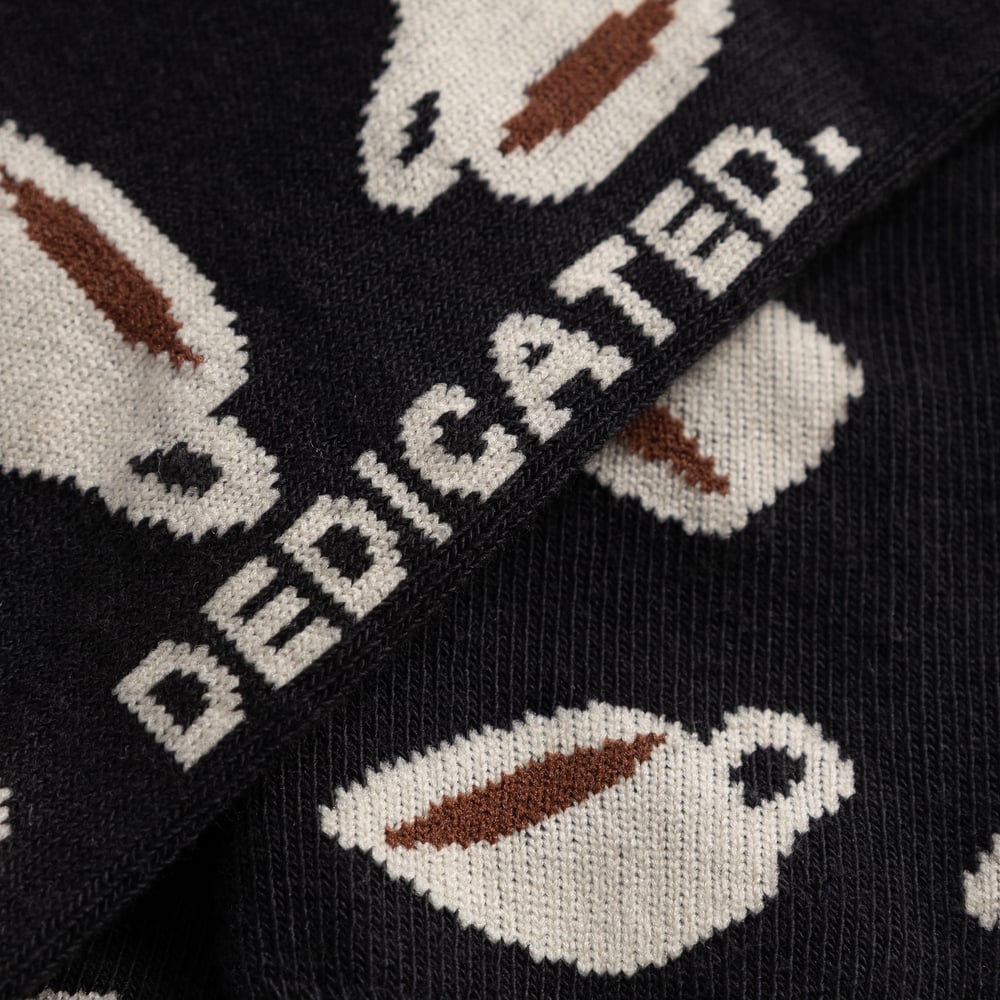 Close-up of the Sigtuna socks in black, showing the detailed coffee cup pattern and the knitted DEDICATED. logo.
