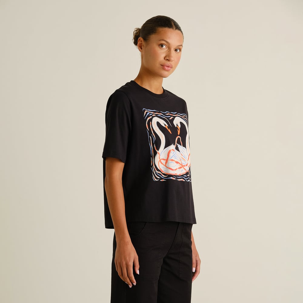 A woman wears the relaxed-fit T-shirt Vadstena in black, featuring a beautiful artistic graphic of two swans.