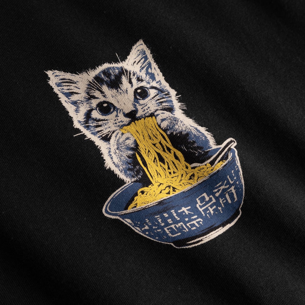Close-up of the playful Cat and Noodles graphic on a black t-shirt, showing a kitten enjoying a big bowl of noodles.