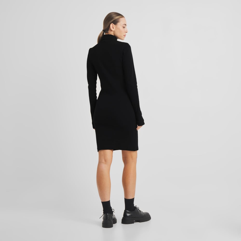 Frau von hinten, die das schwarze langärmelige Rippkleid Lunsen und klobige schwarze Schuhe trägt.
