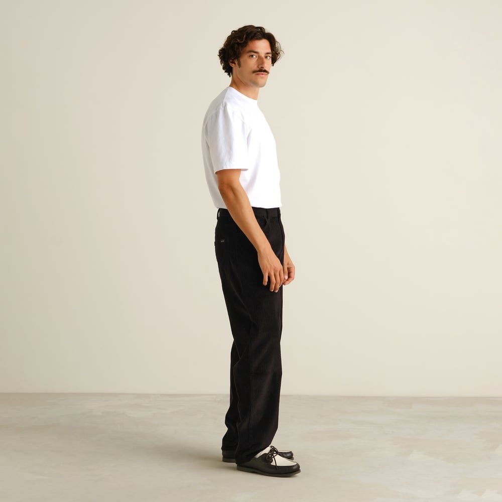 A model in a studio shows off the relaxed fit of the black Hamburg Corduroy Pants, styled with a classic white tee.