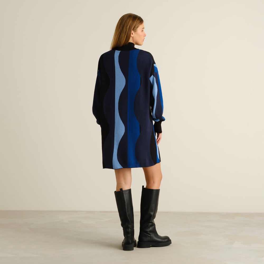Back view of a model wearing the Nikkala dress in a blue wavy stripe pattern, styled with black knee-high boots.