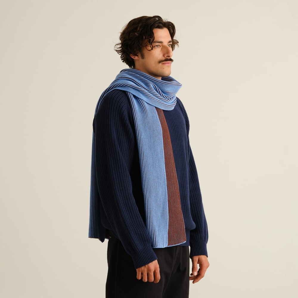 Man i profil bär den randiga halsduken Norrfors i Cappuccino Brown och Cornflower Blue till en marinblå stickad tröja.