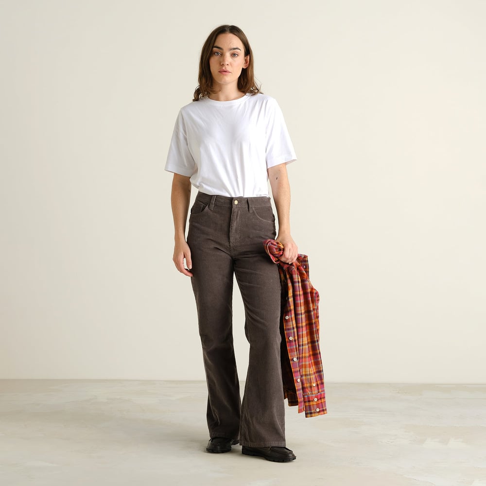 Eine Frau stylt die graue Ryda Bootcut-Cordhose mit einem weißen T-Shirt und hält ein kariertes Hemd in der Hand.