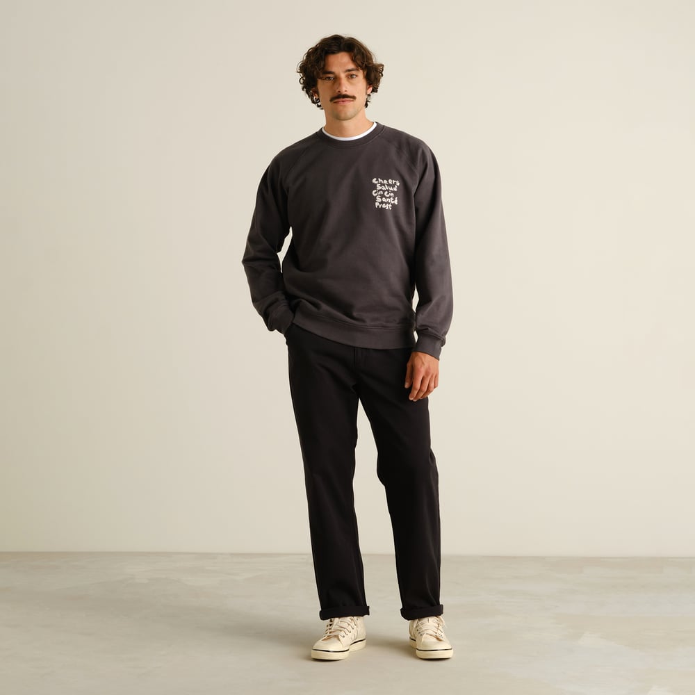 A man with a mustache wearing black Hudiksvall chinos, a dark grey sweatshirt, and off-white sneakers against a background.