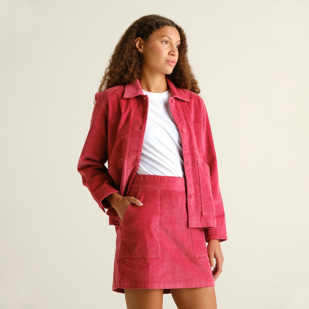 A woman with curly hair models a matching set of a Holly Red corduroy jacket and the Majorna skirt, over a white t-shirt.