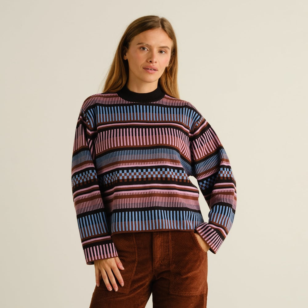 A woman models the Limhamn sweater, featuring a multi-colored dashed stripe pattern, styled with brown corduroy pants.