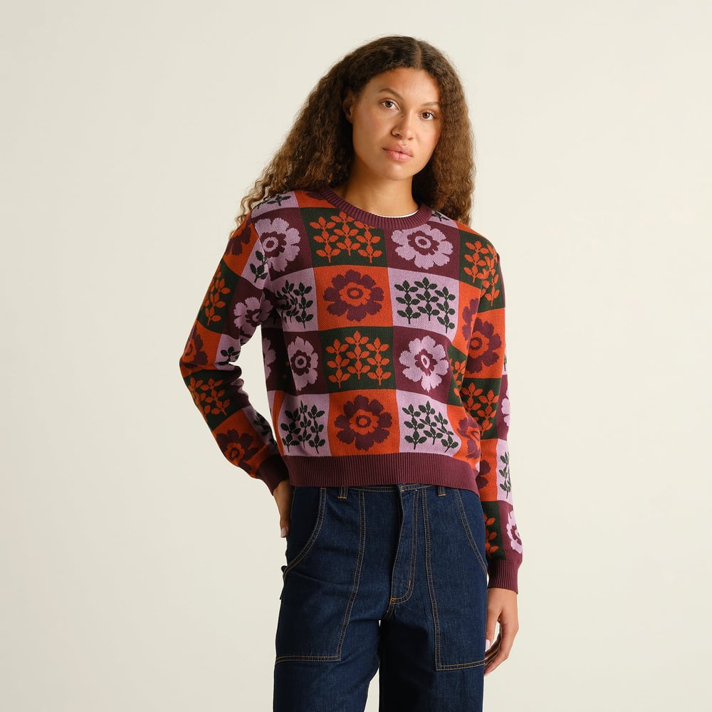 A model wears the jacquard-knit Sweater Arendal, featuring a retro multi-colored floral check pattern with dark wash jeans.