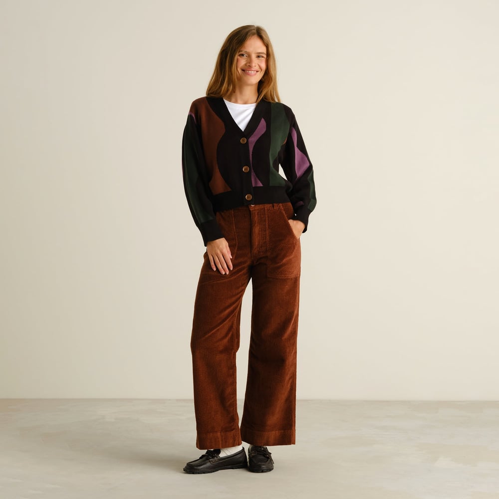 A woman smiling in a studio, wearing the Cardigan Kanan Wavy Stripe with brown corduroy trousers.