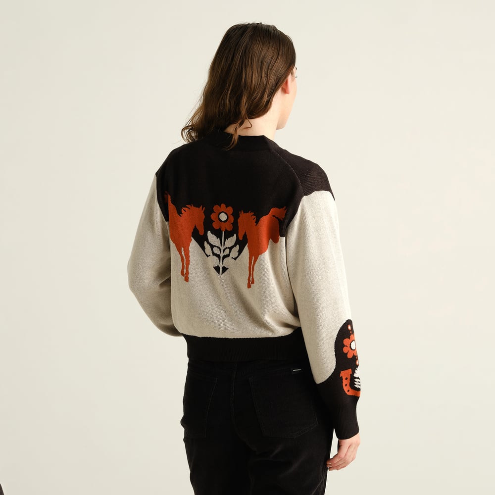A woman seen from the back wears the Kanan Western cardigan, showing off its intricate multi-color horse and flower design.