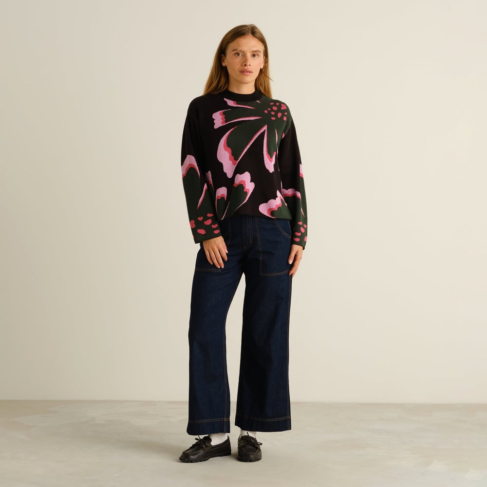 A woman looks straight ahead, wearing the Sweater Limhamn Big Bloom with dark, wide-leg jeans and black loafers.