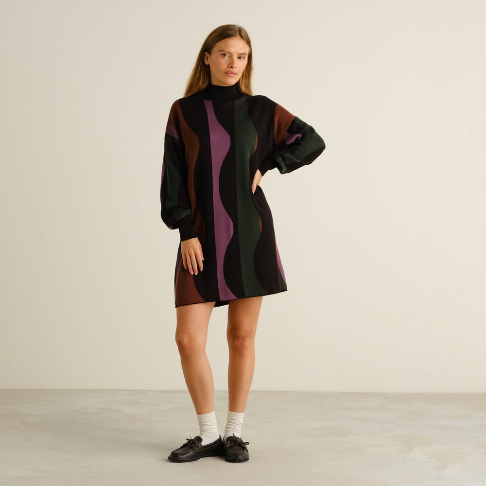 Model looking at the camera wearing Dress Nikkala, a knitted turtleneck dress with a fun multi-colored wavy stripe pattern.