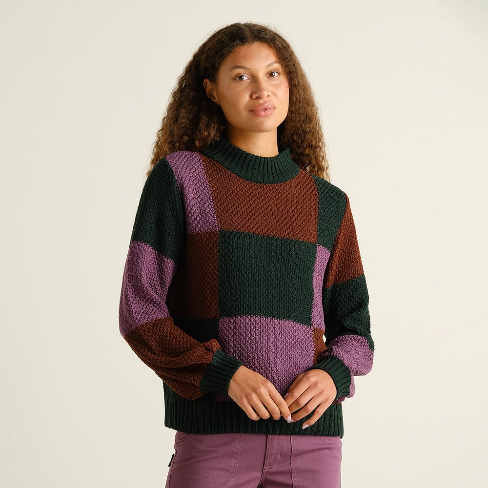 Model wears the chunky-knit Sweater Gotland, featuring a bold blocked check pattern in purple, green, and brown tones.