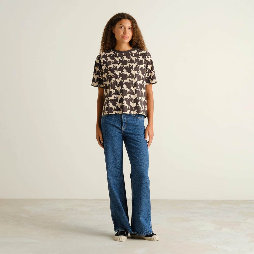 Model with curly hair showcases the T-shirt Vadstena Wild Horses AOP Charcoal, styled with relaxed-fit blue jeans.