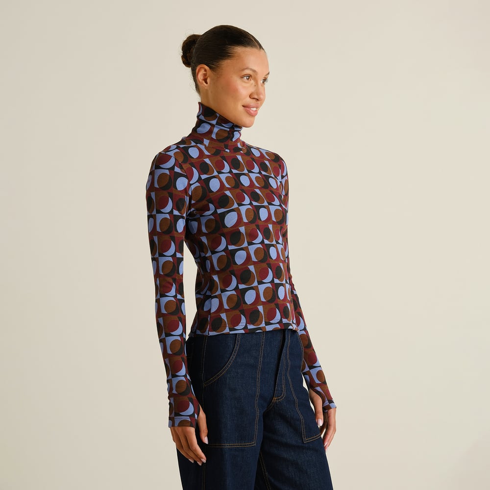 A smiling woman models the slim-fit Rib Top Malma in a fun Retro Geo Multi Color print, paired with dark wash jeans.