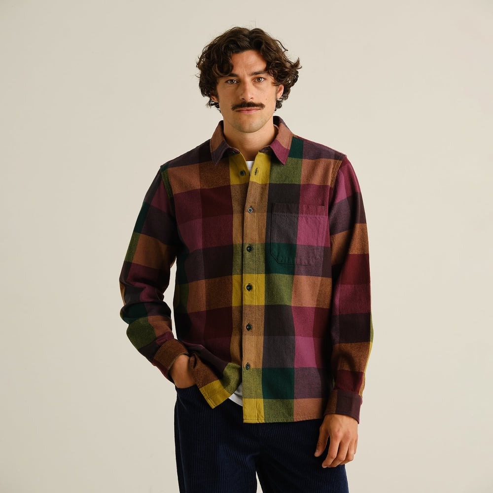 A model faces forward wearing the Dedicated Rute Flannel Shirt in a bold multicolor check with dark corduroy trousers.