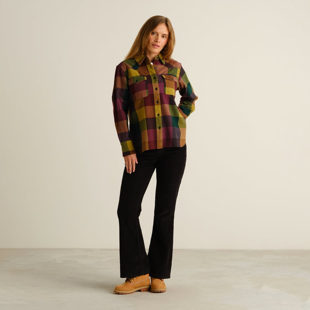 A woman models the western-inspired Arboga Flannel Shirt in a bold multicolor check with black corduroy pants and tan boots.