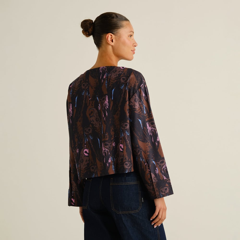 A woman with her hair in a bun shows the back of the Top Udda, a boxy blouse with a vibrant owl print.