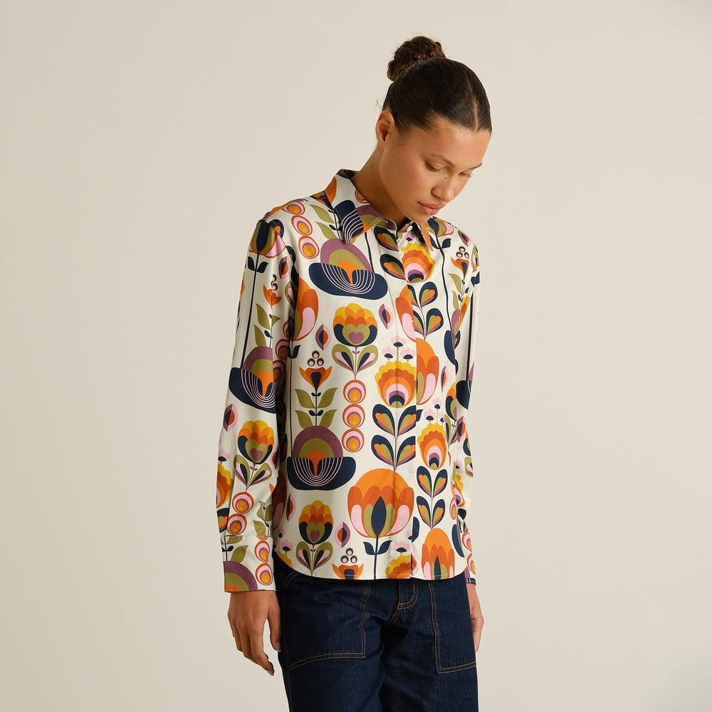 Kvinna i en färgglad, blommönstrad skjorta i retrostil och mörka jeans, med håret uppsatt i en knut.
