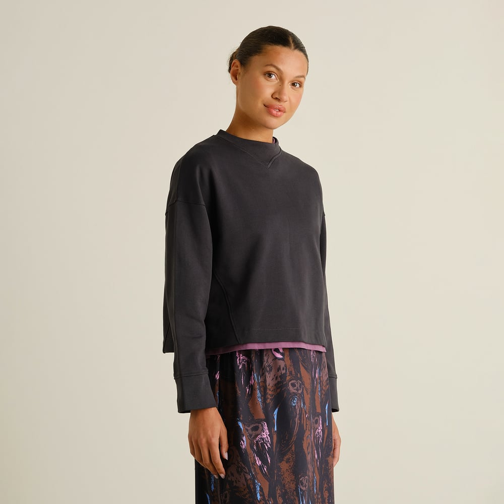 A woman models the loose-fit Sweatshirt Gia in Phantom Black against a plain background, paired with a patterned skirt.