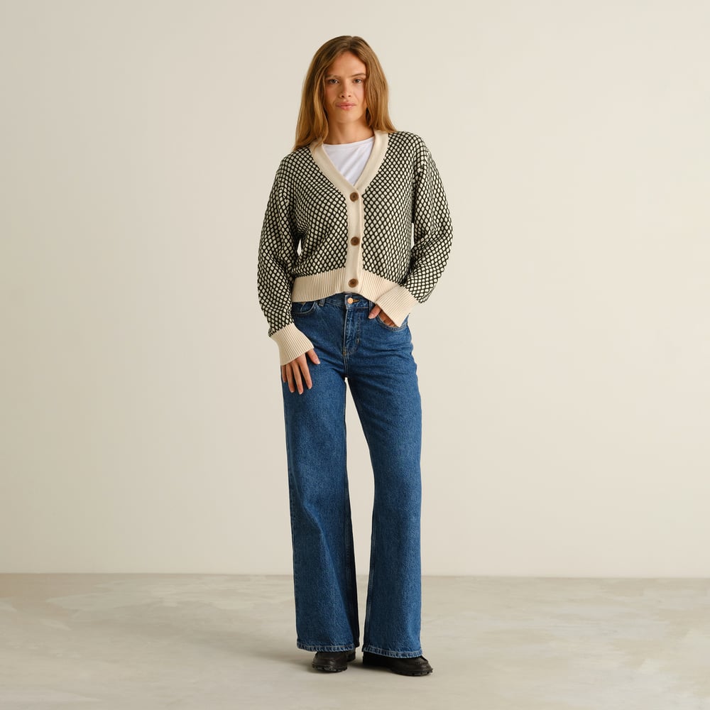 Helkroppsbild av en kvinna som bär den mörkgröna och vaniljfärgade stickade koftan Kanan Bubble Knit med jeans med vida ben.