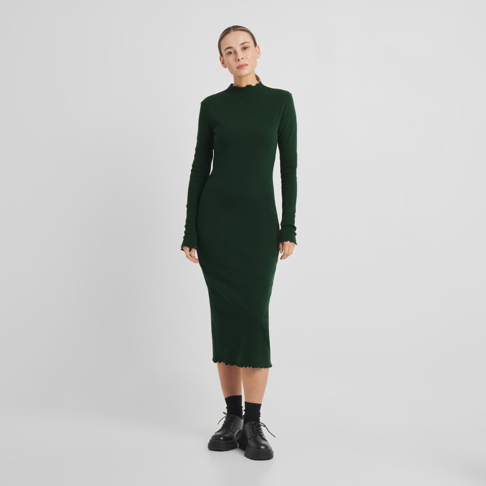 Model wearing the long-sleeved Rib Dress Lofta in dark green, styled with black socks and chunky black shoes.