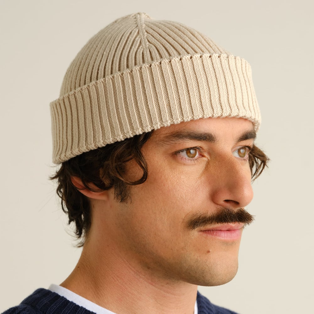 Close-up of a man with a mustache wearing the rib-knit Rutvik Beanie in a soft oat white.