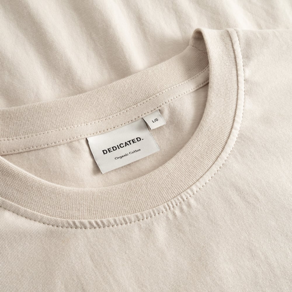 Close-up of the Dedicated brand label and ribbed collar on the classic Stockholm t-shirt in a soft oat white.
