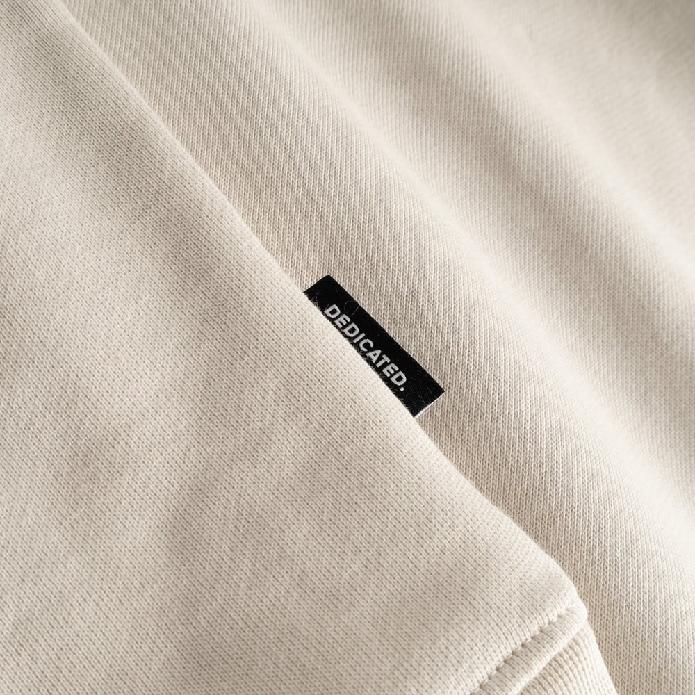 Close-up detail of the signature Dedicated brand label sewn onto the soft, oat-white fabric of a classic sweatshirt.