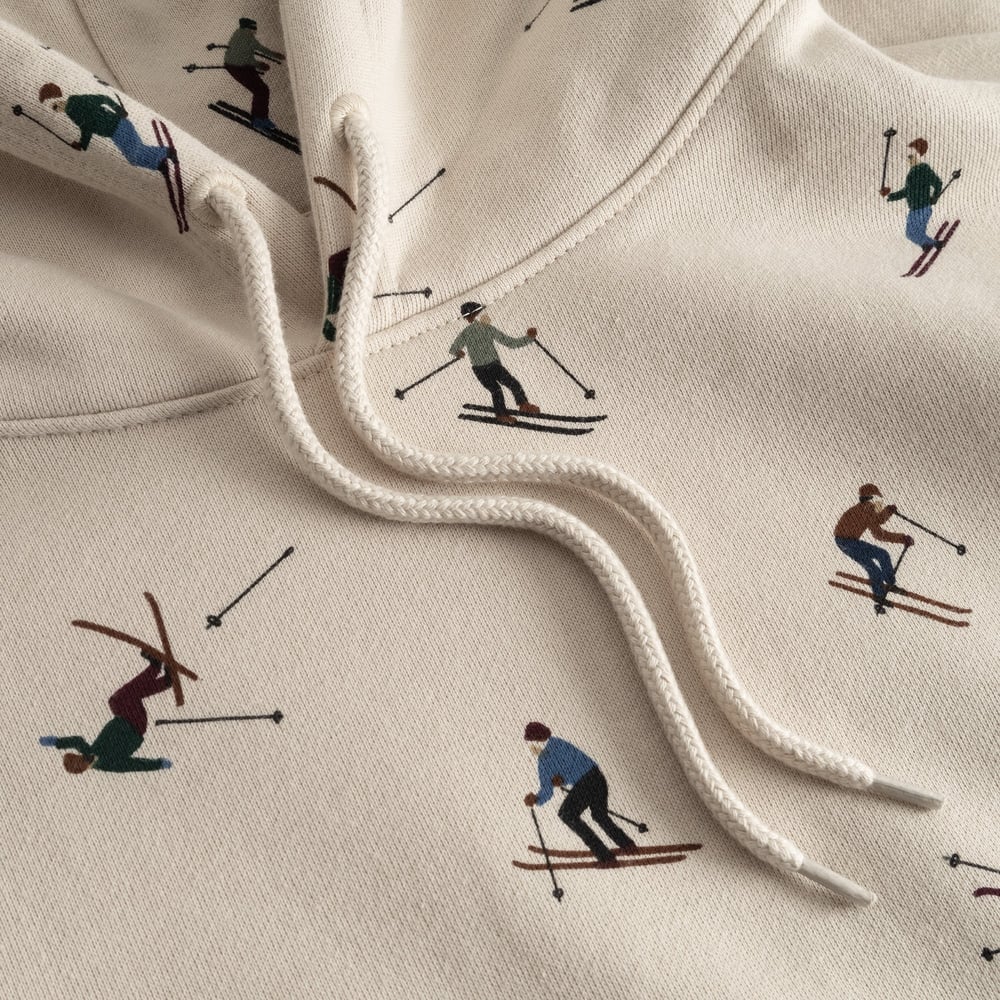 Close-up detail of the oat white Falun Ski Team hoodie, showing the playful all-over embroidered skier print and drawstrings.