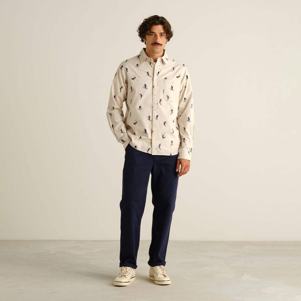 Full-length photo of a model wearing the oat white Shirt Lunnevik Ski Team with dark blue trousers and off-white trainers.
