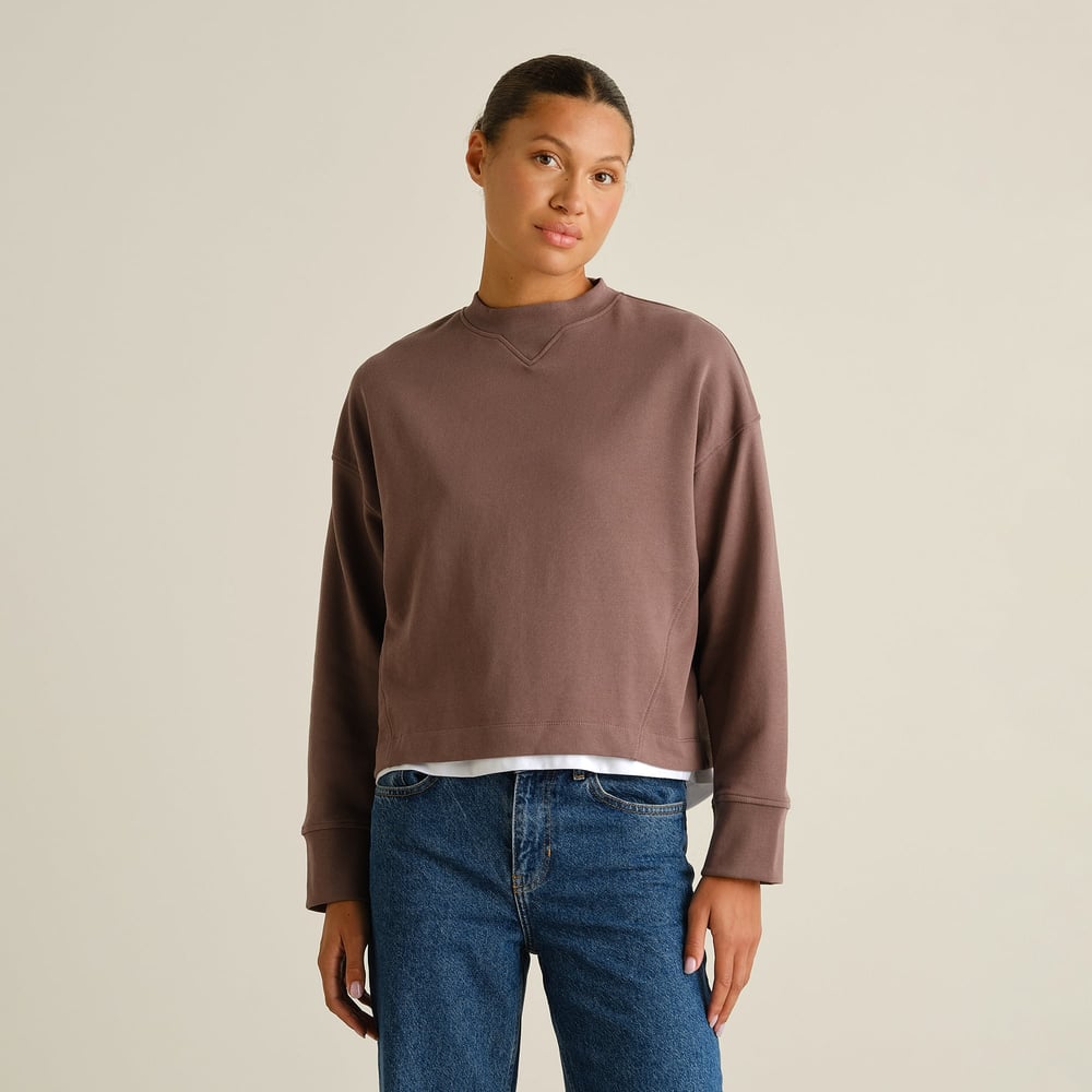 A model wears the Sweatshirt Gia in Sparrow Brown, styled simply with a white t-shirt and classic blue jeans.