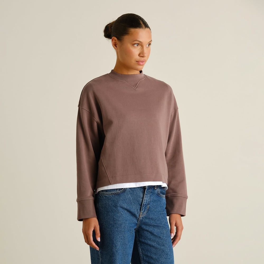 Side view of a woman wearing the Sweatshirt Gia Sparrow Brown, styled with blue jeans and a white tee underneath.