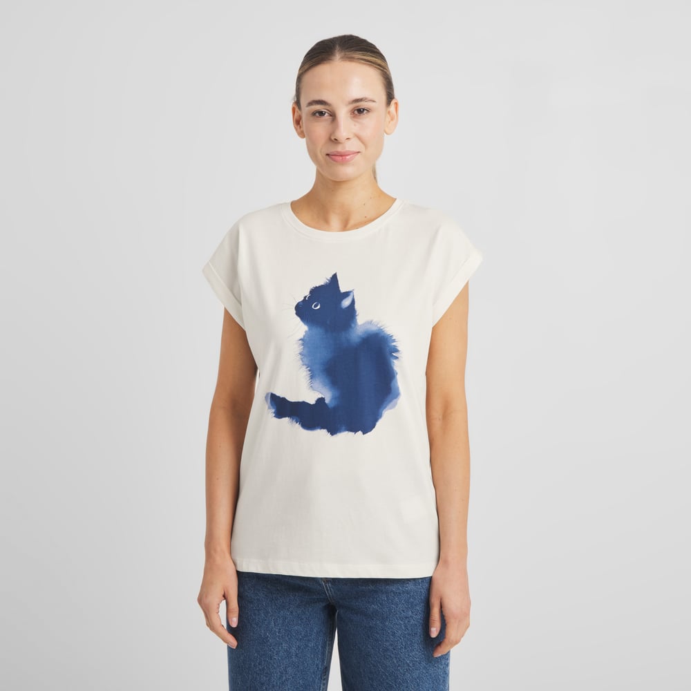 A woman wears the off-white T-shirt Visby, which features a charming blue watercolor cat print, styled with blue jeans.