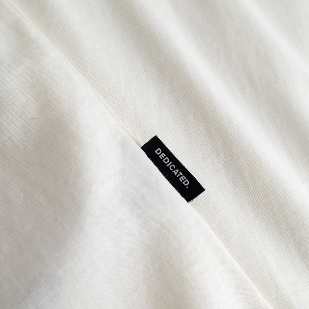 Close-up of the black Dedicated brand tab on the side seam of the off-white T-shirt Stockholm Camp Crew.