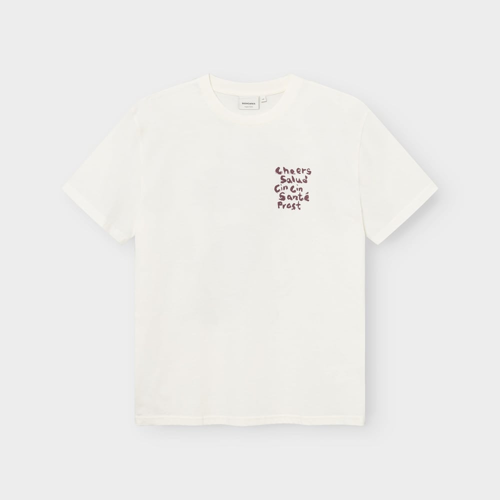 Studioaufnahme des T-Shirts Stockholm Cheers Salud in Cremeweiß mit dem Brustaufdruck „Cheers“ in verschiedenen Sprachen.