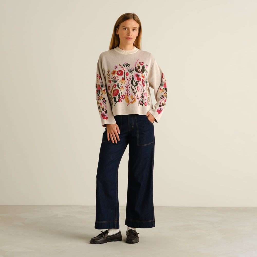 Full-length shot of a model wearing the vanilla white Sweater Limhamn Folk Flower with dark wide-leg jeans and loafers.