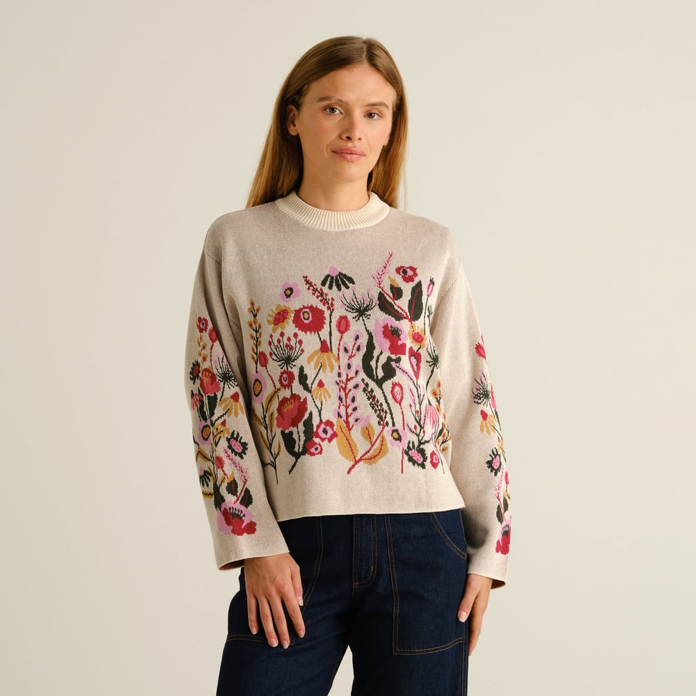 A woman wearing the Limhamn Folk Flower sweater in vanilla white, paired with dark denim jeans, looks towards the camera.