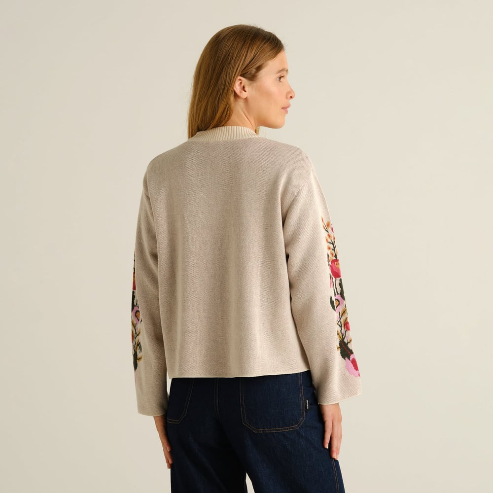 A woman models the back of the vanilla white Limhamn sweater, featuring a vibrant folk flower pattern on the sleeves.