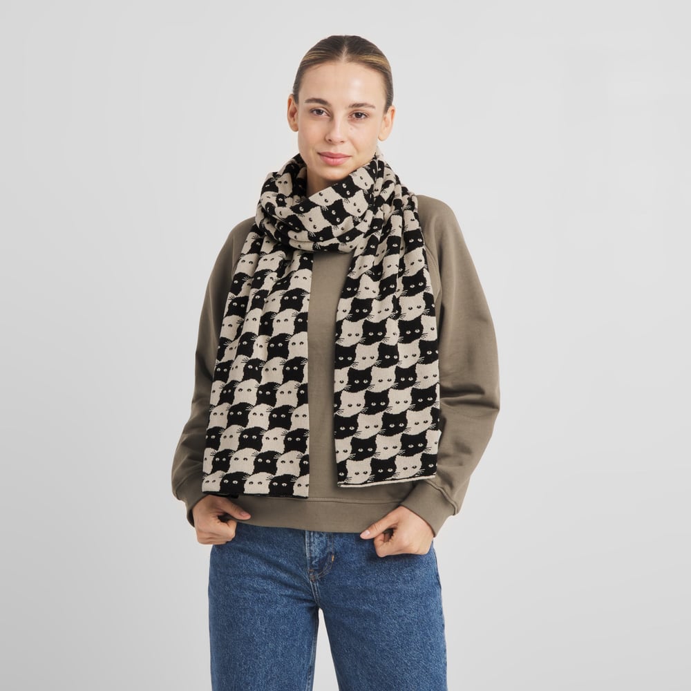 A woman wearing the Scarf Rossberga Pepita Cats in oat white over a beige sweatshirt and blue jeans.