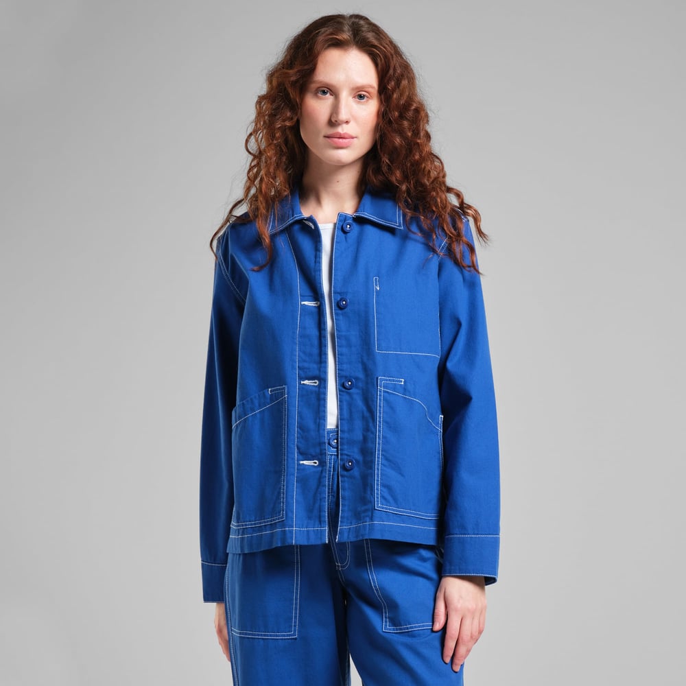 A woman with long, curly hair models the blue Stiby Canvas Jacket, showing its relaxed fit and white contrast stitching.