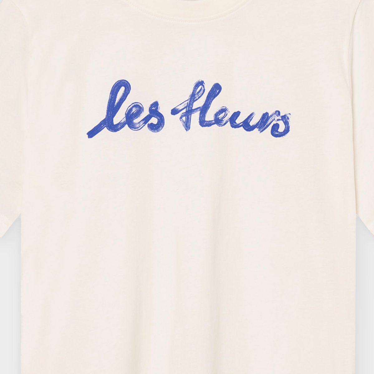 Close-up on the chest of the off-white T-shirt Vadstena, featuring the 'les fleurs' text in a cobalt blue brushstroke script.