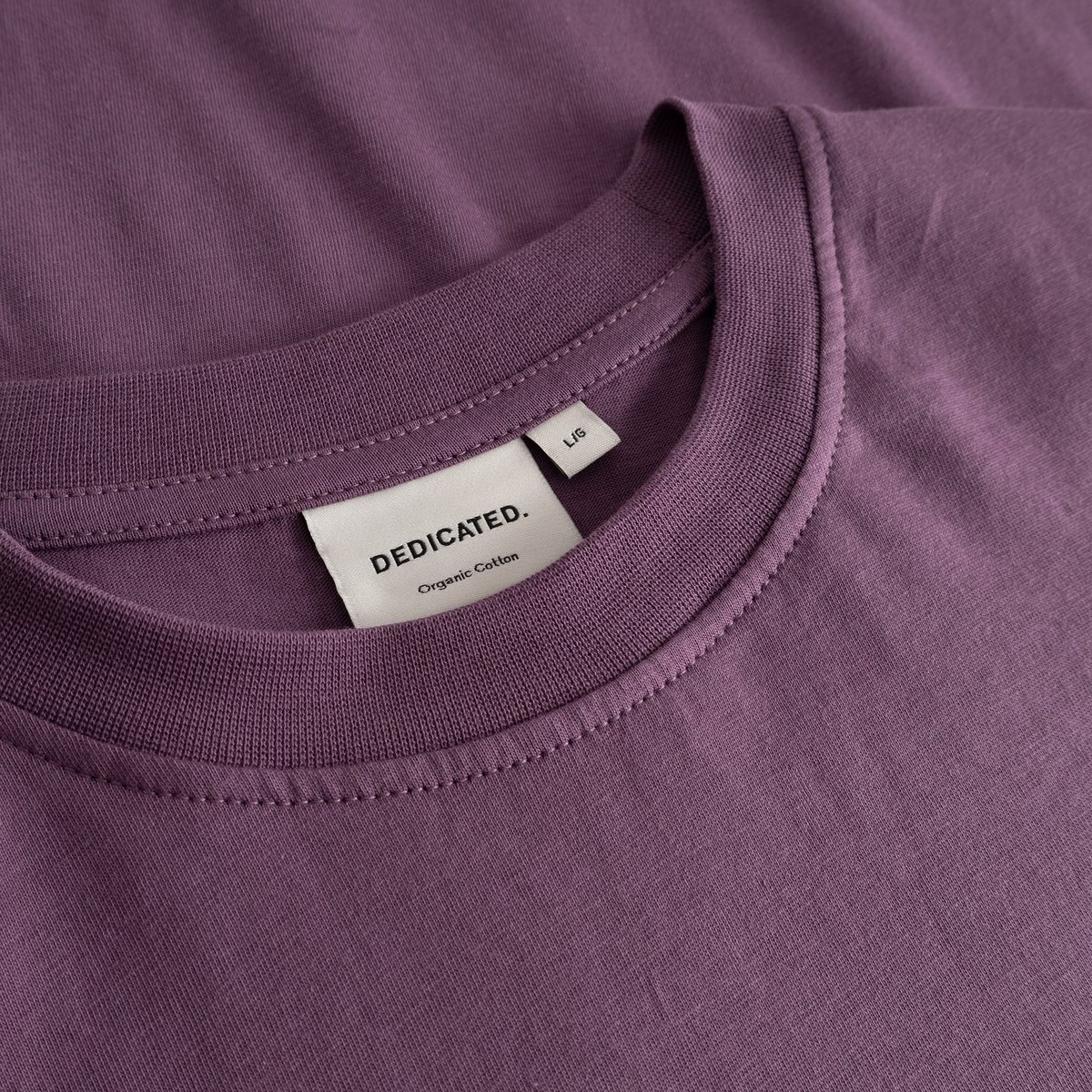Nahaufnahme des Rippkragens und des Dedicated-Etiketts am T-Shirt Stockholm in Berry Purple aus Bio-Baumwolle.