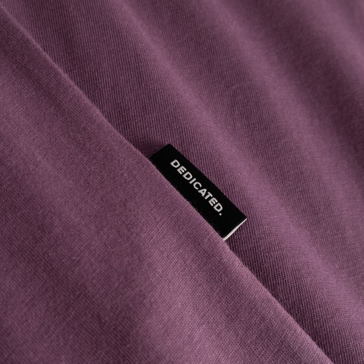 Närbild på T-shirt Stockholm Base i Berry Purple, som visar tygstrukturen och den lilla Dedicated-etiketten.