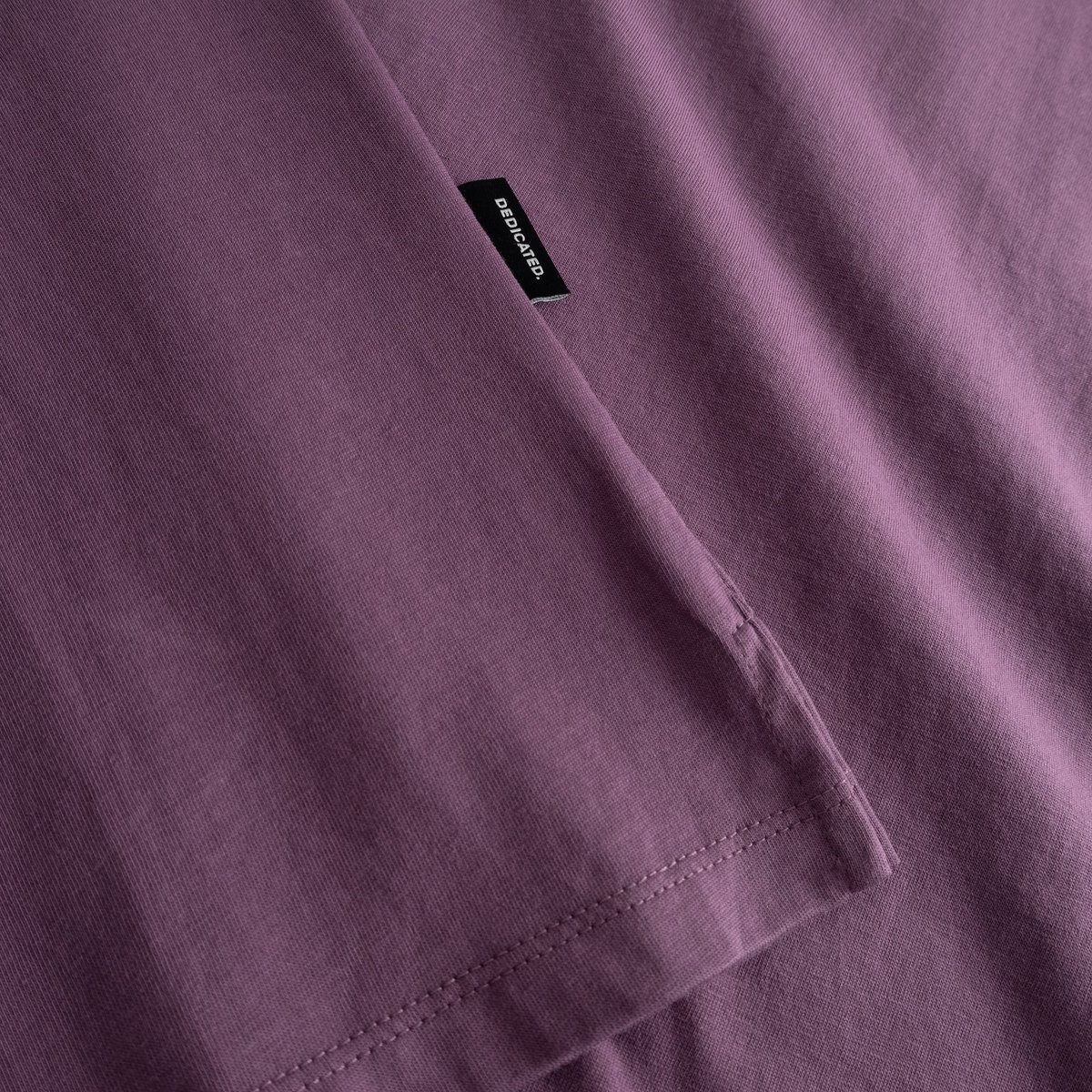 Close-up of the folded sleeve, side slit, and brand tag on the T-shirt Visby Base in a rich Berry Purple hue.