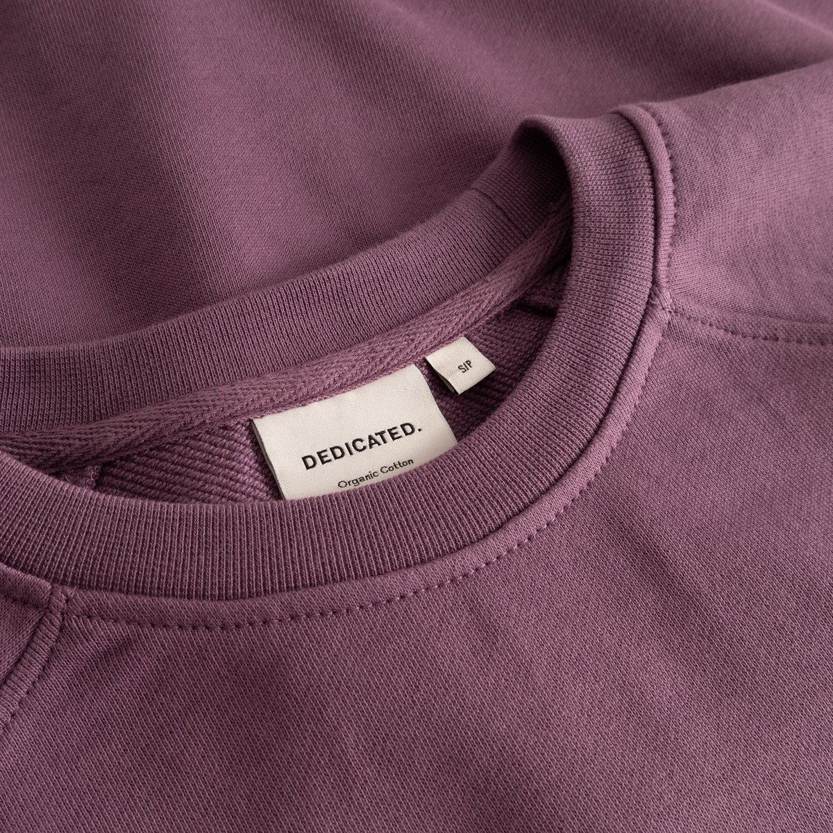 A close-up of the Ystad Sweatshirt in Berry Purple, detailing the soft ribbed crew neck and the classic Dedicated brand tag.