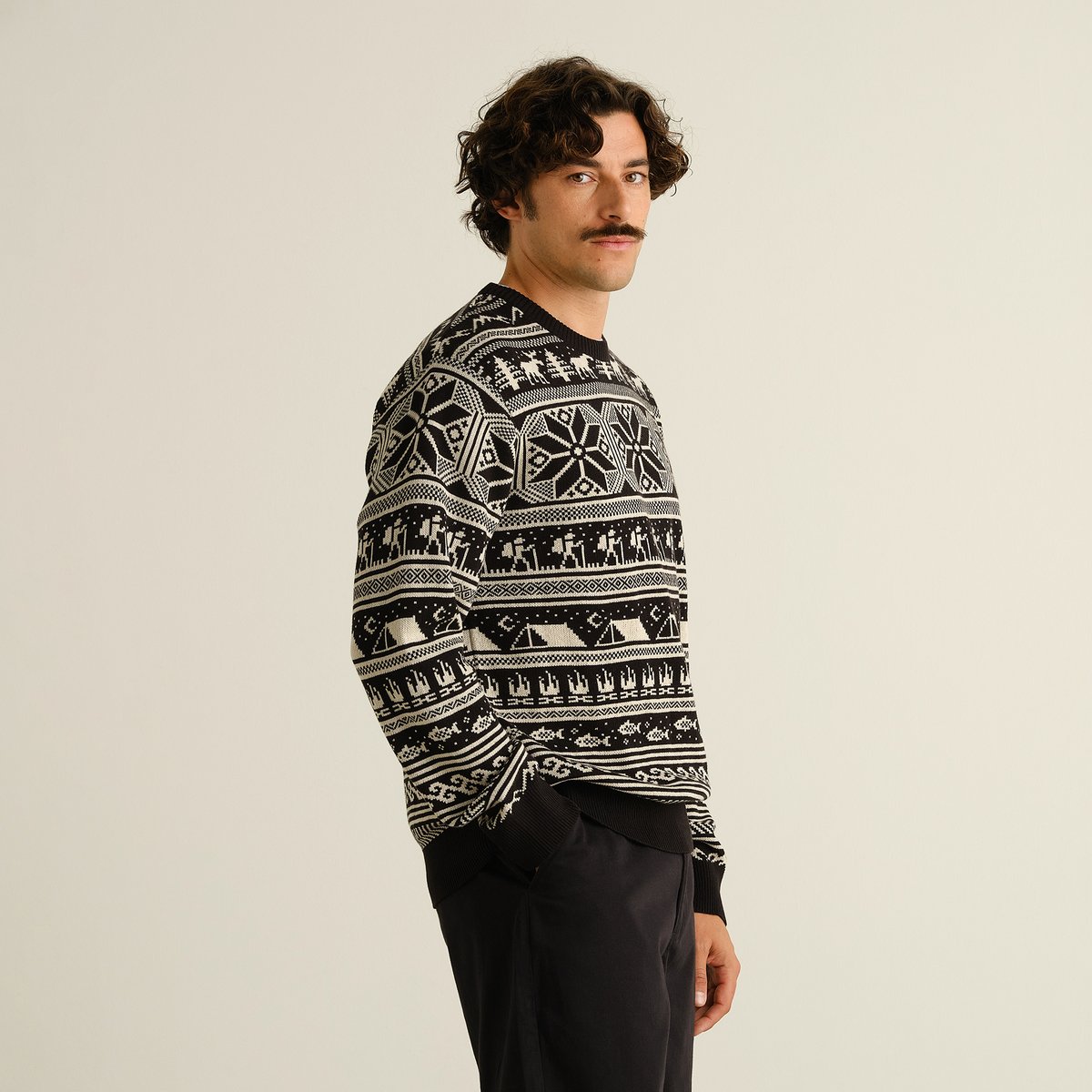 Side view of a model wearing the black Sweater Mora Fairisle Hike, featuring a pattern of hikers, tents, and snowflakes.