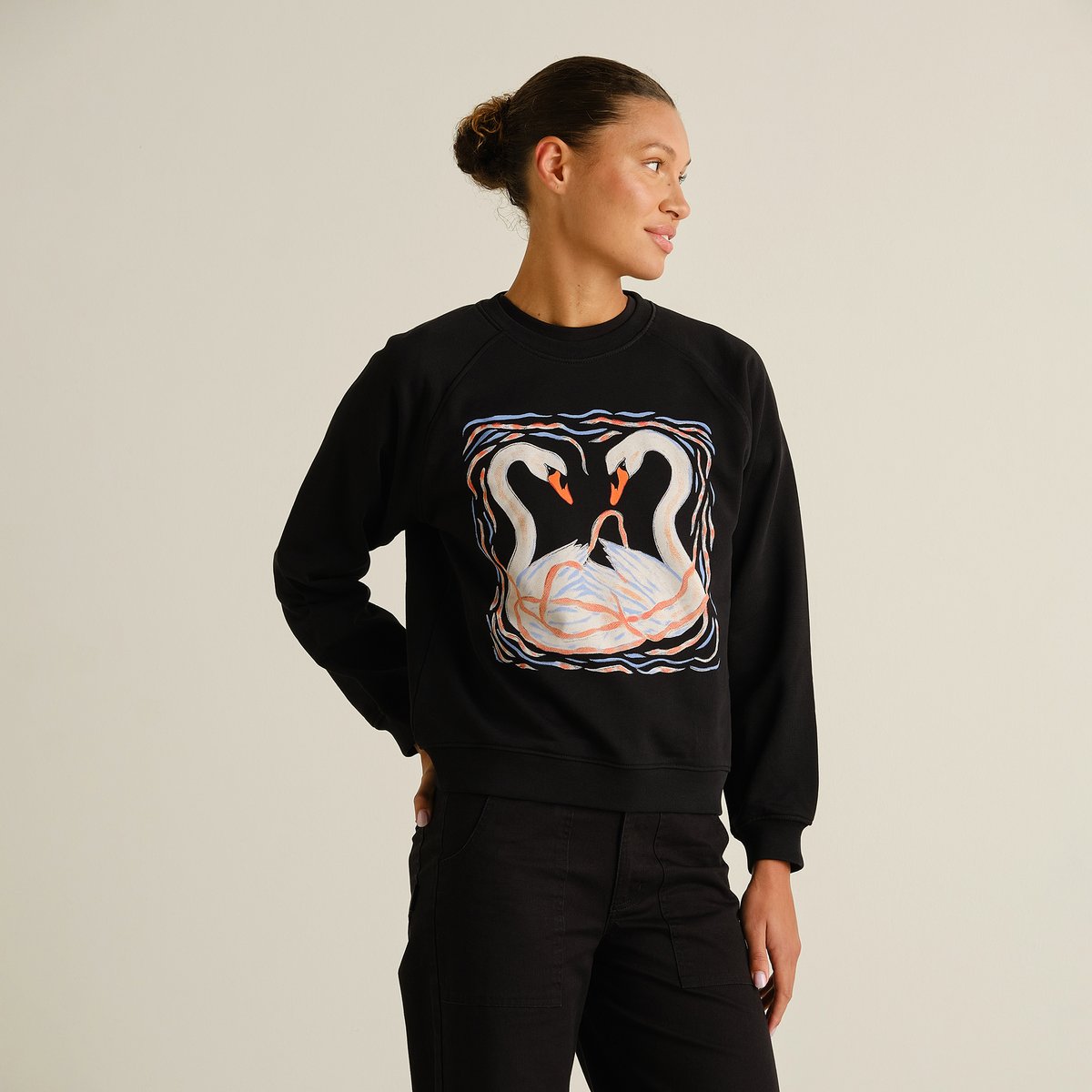 A woman models the black Ystad sweatshirt with a painted swans graphic, looking over her shoulder against a plain backdrop.
