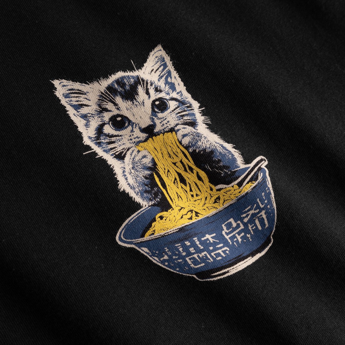 Close-up of the playful Cat and Noodles graphic on a black t-shirt, showing a kitten enjoying a big bowl of noodles.