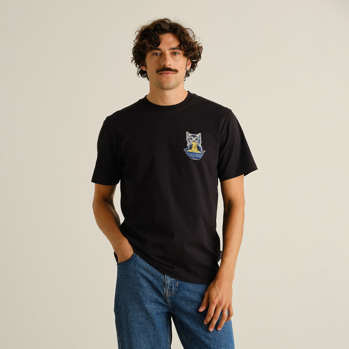 A model wears the black T-shirt Stockholm Cat and Noodles with a chest print and blue jeans against a studio background.